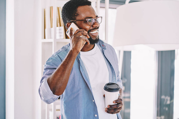 Cheerful african american male in eyewear talking on mobile phone with friend laughing on work break, dark skinned  hipster guy having telephone conversation holding cardboard coffee cup in offic - Photo, Image