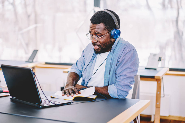 Young african american man in modern headphones listening audio book for education doing homework, dark skinned hipster guy entertaining with playlist on netbook writing ideas for articl - Foto, Imagem