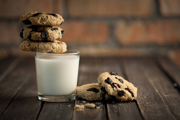 A glass of milk and cookies with raisins. A wooden table and a brick wall on a background. Selective focus. - Фото, изображение
