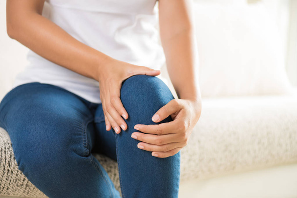 Nahaufnahme junge Frau sitzt auf dem Sofa und spürt Knieschmerzen und sie massiert ihr Knie zu Hause. Gesundheitswesen und medizinisches Konzept. - Foto, Bild