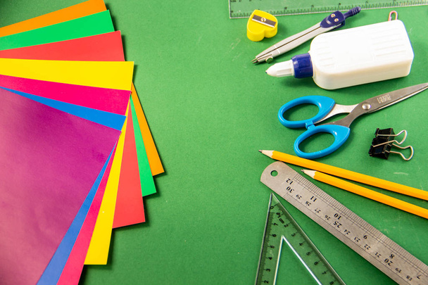 Pennen, potloden, paperclips, kleurpotloden, linialen, lijm, corrector marker en gekleurd papier op een groene achtergrond voelde - Foto, afbeelding