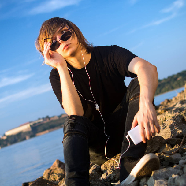 A tizenéves fiú lóg, egyedül a folyóparti és hallás zene-ból mozgatható telefon. - Fotó, kép