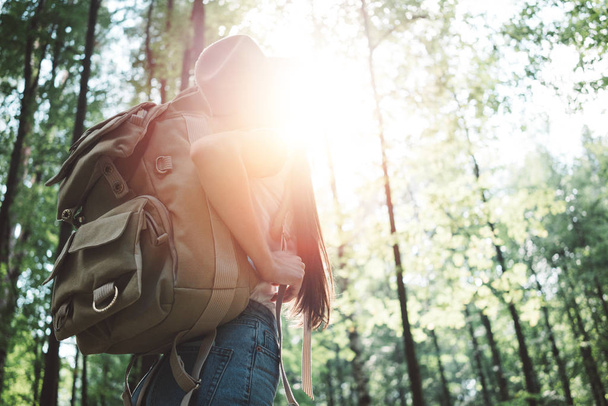 Handsome traveler woman with backpack and hat standing in forest. Young hipster girl walking among trees on sunset - Foto, imagen