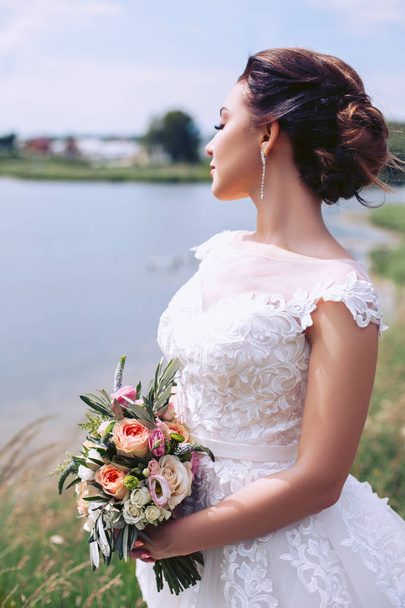 Young bride in a wedding dress is standing on the river bank with a bouquet of flowers - Фото, изображение