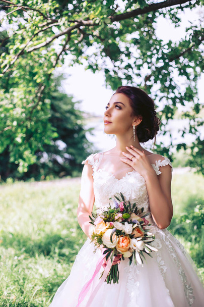 A young bride on a sunny day stands under a tree with a bouquet of flowers - Foto, Imagen