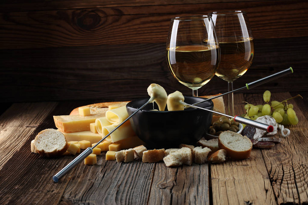 Gourmet Swiss fondue dinner on a winter evening with assorted cheeses on a board alongside a heated pot of cheese fondue with two forks dipping bread and white wine behind in a tavern or restaurant - Фото, изображение