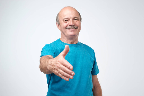 You are welcome concept. Cheerful mature man in blue t-shirt gesturing welcome sign and smiling while standing against gray wall. He is happy to meet dear guests - Photo, Image