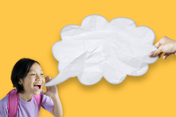 Portrait of Asian schoolgirl looks happy while shouting at empty cloud bubble through her hand. Shot with yellow screen - Photo, image