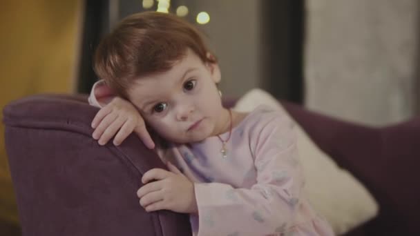 little girl is sitting on the couch and is looking at the camera - Кадри, відео