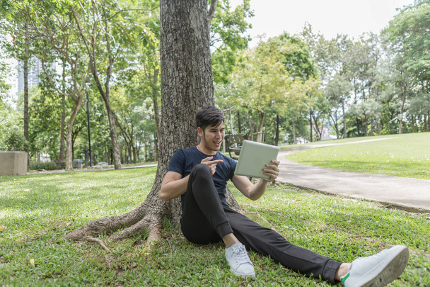 Nuori liikemies käyttää tablettia ja nojaa puuhun puistossa
. - Valokuva, kuva