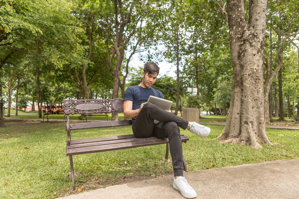 junger Geschäftsmann bedient sich des Tablets und setzt sich im Park auf den Stuhl. - Foto, Bild