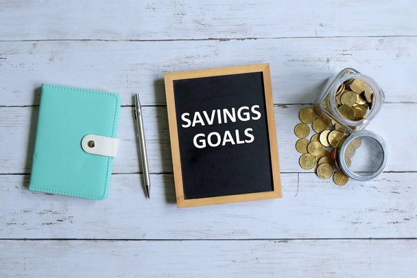 ノート、ペン、コインと白い木製の背景に '貯蓄目標' と書かれた黒板の上から見る. - 写真・画像