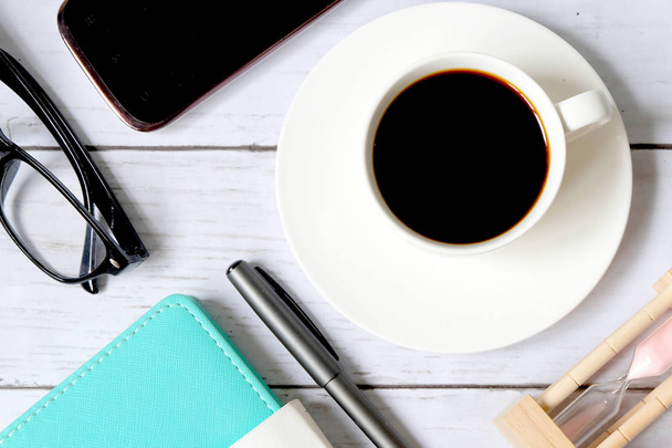 Top view and selective focus of a cup of black coffee with hourglass,sunglasses, notebook,mobile phone and pen on white wooden background. - 写真・画像