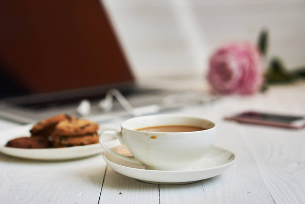 Hot cup of fresh coffee and laptop on the work desk. Cosy home and work concept - vintage wooden table with laptop computer, coffee cup and biscuit. Women's elegant desktop. Selective focus. - 写真・画像
