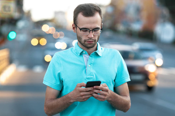 Junger Mann mit Brille benutzt Telefon auf dem Hintergrund der Straße bei Sonnenuntergang. Lifestylekonzept - Foto, Bild