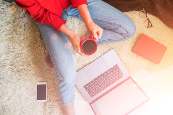 Top view of young woman sitting on floor with laptop. Young woman dressed in red pullover sitting on floor and holding laptop while using smartphone. Online shopping. Girl chooses gifts online. - Foto, immagini