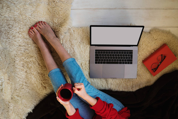 Top view of young woman sitting on floor with laptop. Young woman dressed in red pullover sitting on floor and holding laptop while using smartphone. Online shopping. Girl chooses gifts online. - Foto, afbeelding