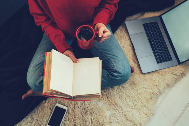 Νεαρή γυναίκα κάθεται στο λευκό χαλί με βιβλία, smartphone και laptop στο σύγχρονο σαλόνι, μελετώντας και χαλαρώνετε στο σπίτι. Άνετο εσωτερικό και κορίτσι, διαβάζοντας ένα βιβλίο, οι έννοιες του σπιτιού και άνεση. - Φωτογραφία, εικόνα
