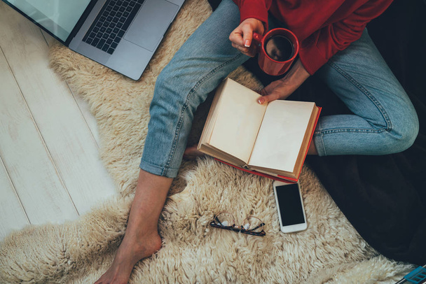 Young woman sitting on white carpet with books, smartphone and laptop in modern living room, studying and relaxing at home. Cozy interior and girl, reading a book, concepts of home and comfort. - Φωτογραφία, εικόνα