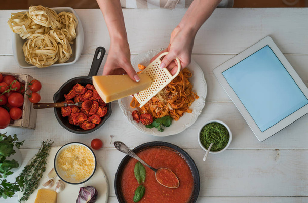 Vrouw in de keuken bereiden van lekkere, zelfgemaakte pasta in gezellige keuken. Vrouwelijke handen kaas raspen. Pasta met pestosaus, basilicum, kers-tomaten en knoflook op oude grunge witte tafel, bovenaanzicht. - Foto, afbeelding