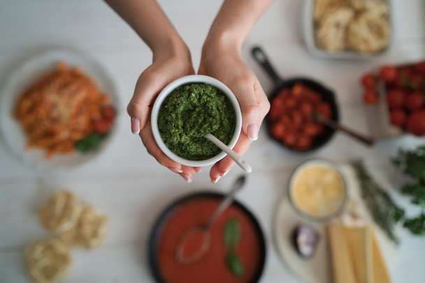 Woman holding small bowl with fresh homemade pesto. Cooking at home. Healthy organic food concept. Vegetarian vegetables, organic food, raw ingredient. Natural nutrition diet. Toned picture. - Photo, Image
