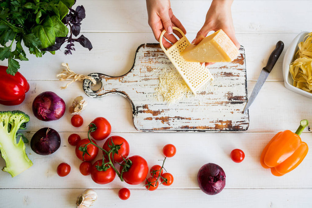 weibliche Hand geschnittenen Käse auf weißen ländlichen hölzernen Küchentisch mit Gemüse Kochzutaten und Werkzeuge, Draufsicht. Lebensmittel-Hintergrund. Food-Konzept mit verschiedenen frischen Zutaten. - Foto, Bild