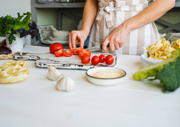 Mujer joven en delantal en la mesa con verduras. buscando comida en casa, preparando el almuerzo de verduras orgánicas frescas en cocina ligera con superficie de madera, llena de utensilios de cocina rurales
.  - Foto, imagen