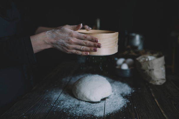 Woman sprinkling flour over fresh dough on kitchen table. Woman is cooking Easter cross buns in a home kitchen. Fresh raw dough for pizza or bread baking on wooden board on kitchen background. - Fotó, kép