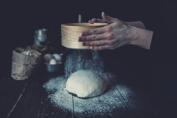 Woman sift the flour through a sieve to prepare the dough for pizza on a dark background. Sprinkling of dough with white wheat flour. Low key shot, close up on hands, some ingredients on table. - Foto, immagini