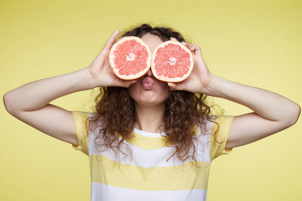 Isolated portrait of young curly haired female holding halved oranges at eyes. Headshot of funny girl wearing stripe T-shirt poses on yellow wall, sending kiss to boyfriend. Expressions and emotions.  - Photo, Image