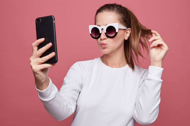Shot of funny female model in stylish eyewear, poses at camera of smart phone, makes grimace, holds modern smart phone. Beautiful woman uses gadget for taking photo. Youth and technology concept. - Foto, Bild