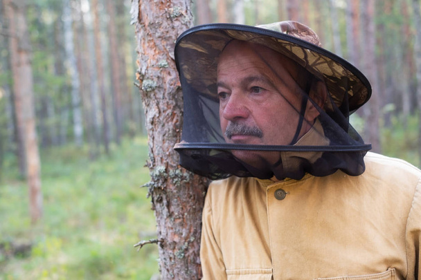 Älterer kaukasischer Mann mit spezieller Mückenschutzmütze, der im Wald steht und sich ausruht, während er Pilze sammelt - Foto, Bild
