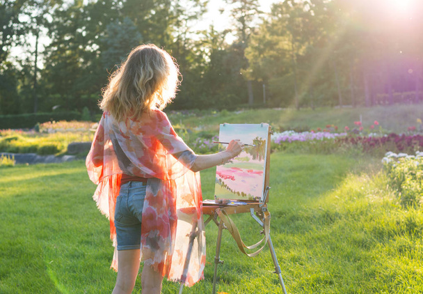 Una joven artista sostiene un pincel y pinta un cuadro en un caballete en los rayos de la puesta del sol. El pintor pinta pinturas al óleo en el jardín al atardecer
 - Foto, Imagen
