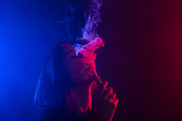 Młoda kobieta w neon light i dym z e papierosy lub vape na ciemnym tle - Zdjęcie, obraz