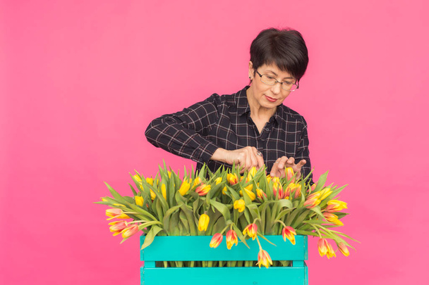 Piękna kobieta w średnim wieku z żółtych tulipanów na różowym tle. Koncepcja florystyki, święta i prezenty - Zdjęcie, obraz