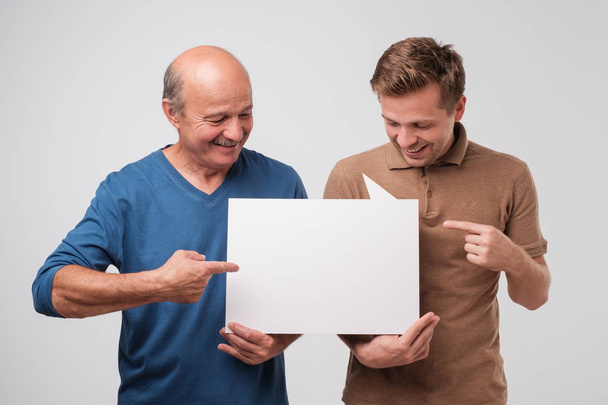 Двое взрослых мужчин отец и сын держат белый пустой рекламный щит вместе. Друзья стоят и улыбаются. Обратите внимание на выгодное предложение
 - Фото, изображение