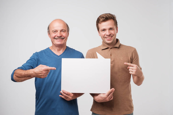 Двое взрослых мужчин отец и сын держат белый пустой рекламный щит вместе. Друзья стоят и улыбаются. Обратите внимание на выгодное предложение
 - Фото, изображение