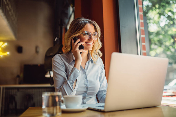 Portrait d'une jeune femme d'affaires avec des lunettes assise dans un café devant son ordinateur portable et parlant sur un téléphone portable
 - Photo, image