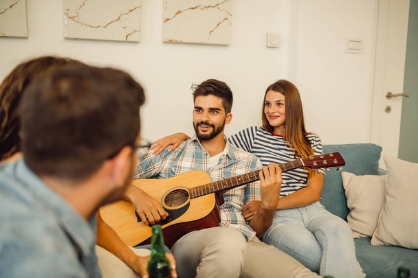 Gruppo di amici sono seduti in casa e divertirsi mentre uno di loro sta suonando la chitarra
. - Foto, immagini