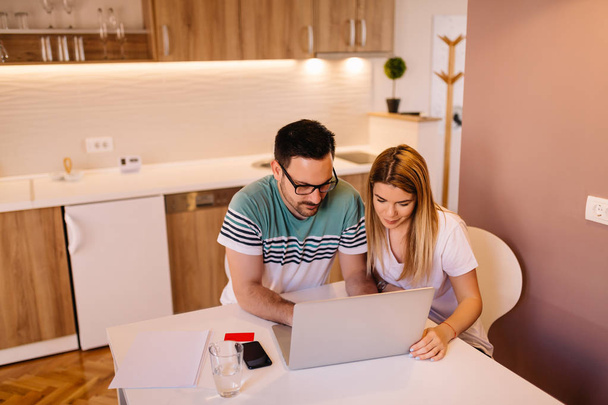 Retrato de jovem casal que está trabalhando em conjunto no laptop enquanto está sentado em uma cozinha moderna em casa
 - Foto, Imagem