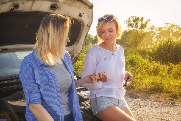 zwei schöne blonde Mädchen haben Spaß bei der Diskussion über Autoreparaturen auf einer Landstraße im Sonnenuntergang. - Foto, Bild