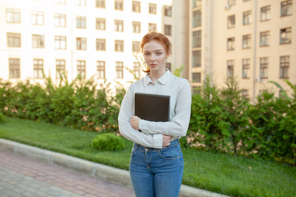 A sad red-haired girl with freckles on her face holds a folder in her hands. Photo of a student before a difficult exam. Back to school concept - Foto, Bild