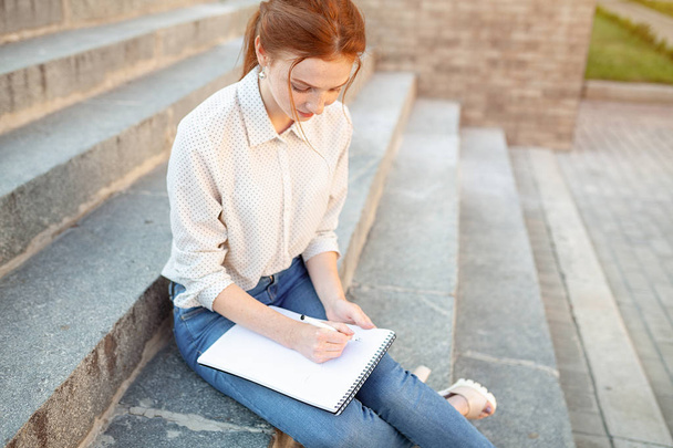 A beautiful young redhaired girl writes a romantic love letter sitting on a stairs near the institute. Student love. Or homework - Foto, afbeelding