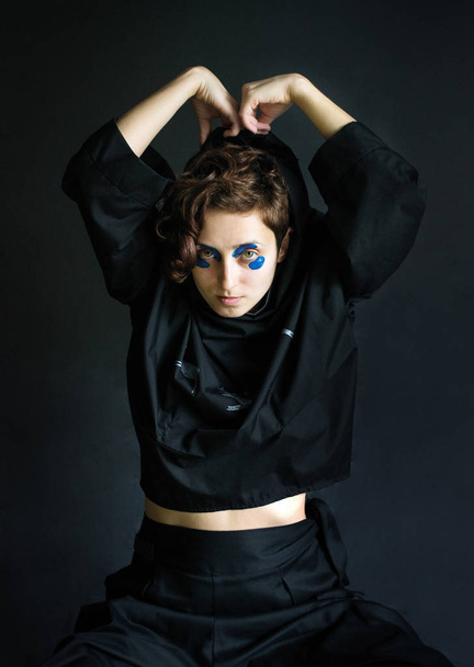 foto Muchacha de fotos de moda con pelo rizado en ropa negra con pintura azul alrededor de sus ojos se quita la ropa
 - Foto, imagen