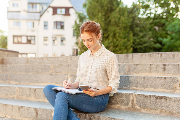 beautiful young redhead girl with freckles sitting on a stairs and making notes in a notebook. Portrait of a student. Remote education. Homework - Foto, Imagen