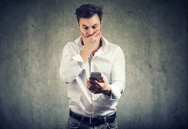 Κομψό νεαρό άνδρα που καλύπτει στόμα σε στρες και έκπληξη βλέποντας smartphone στέκεται σε γκρίζο φόντο - Φωτογραφία, εικόνα