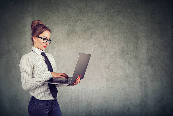 Serious professional woman in formal outfit and glasses using laptop and working against gray wall background - Photo, image