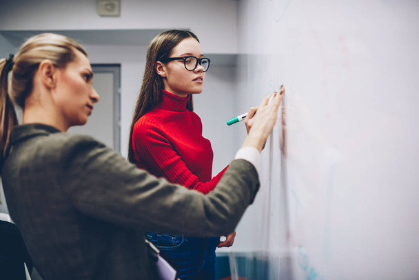 Female colleagues standing near flip chart for discussing strategy and ideas on brainstorming session, skilled woman coach explaining information for young girl student on lesson writing on board - Foto, afbeelding