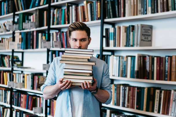 Overworked persistent young man in casual wear holding many literature books in hands for science course work.Intelligent student with textbooks standing in library interior with book shelves - Photo, Image