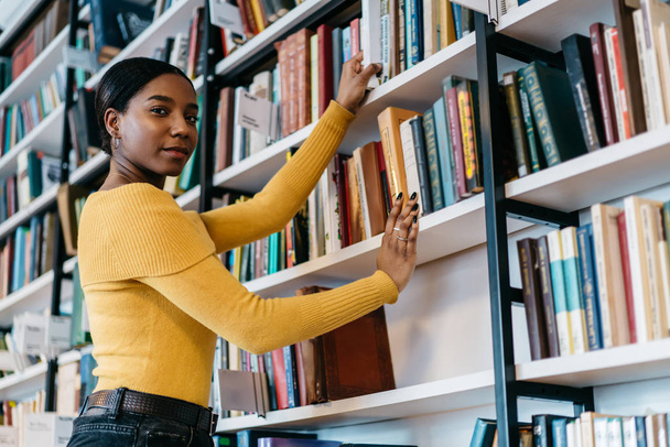 Half length portrait of african american young woman in casual wear looking at camera while pulling literature book from bookshelf in public library.Dark skinned student choosing textbook for studying - Foto, Imagem